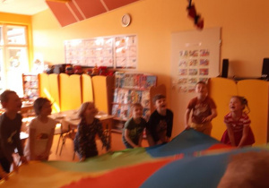 Grupa dzieci faluje chustą animacyjną na której środku leży maskotka Krasnala Hałabały.